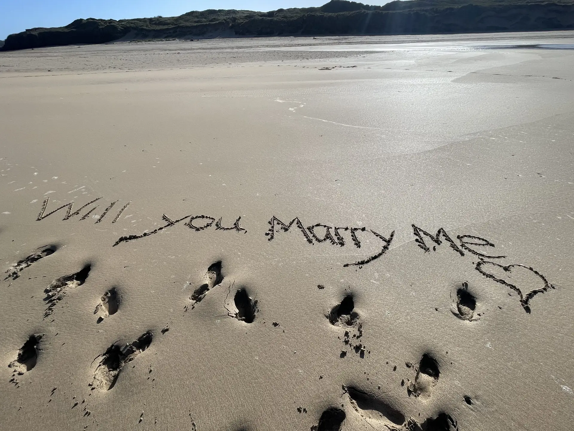 Will You Marry Me Written into the golden sand with a blue sky in the background