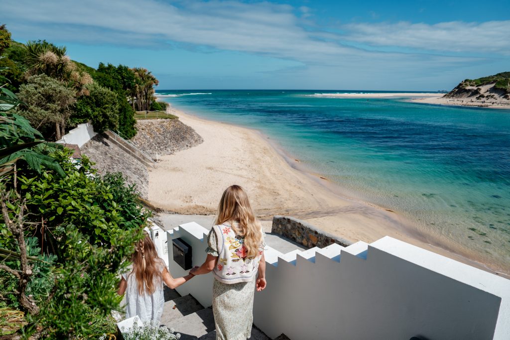 Two people walking down white steps to golden sandy beach and blue sea with blue sky with luscious green gardens
