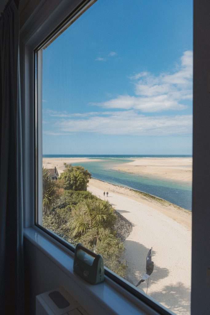 Beach House Bedroom View overlooking the the beach and sea meandering out to a bay with blue sky and white clouds with two people walking on the beach